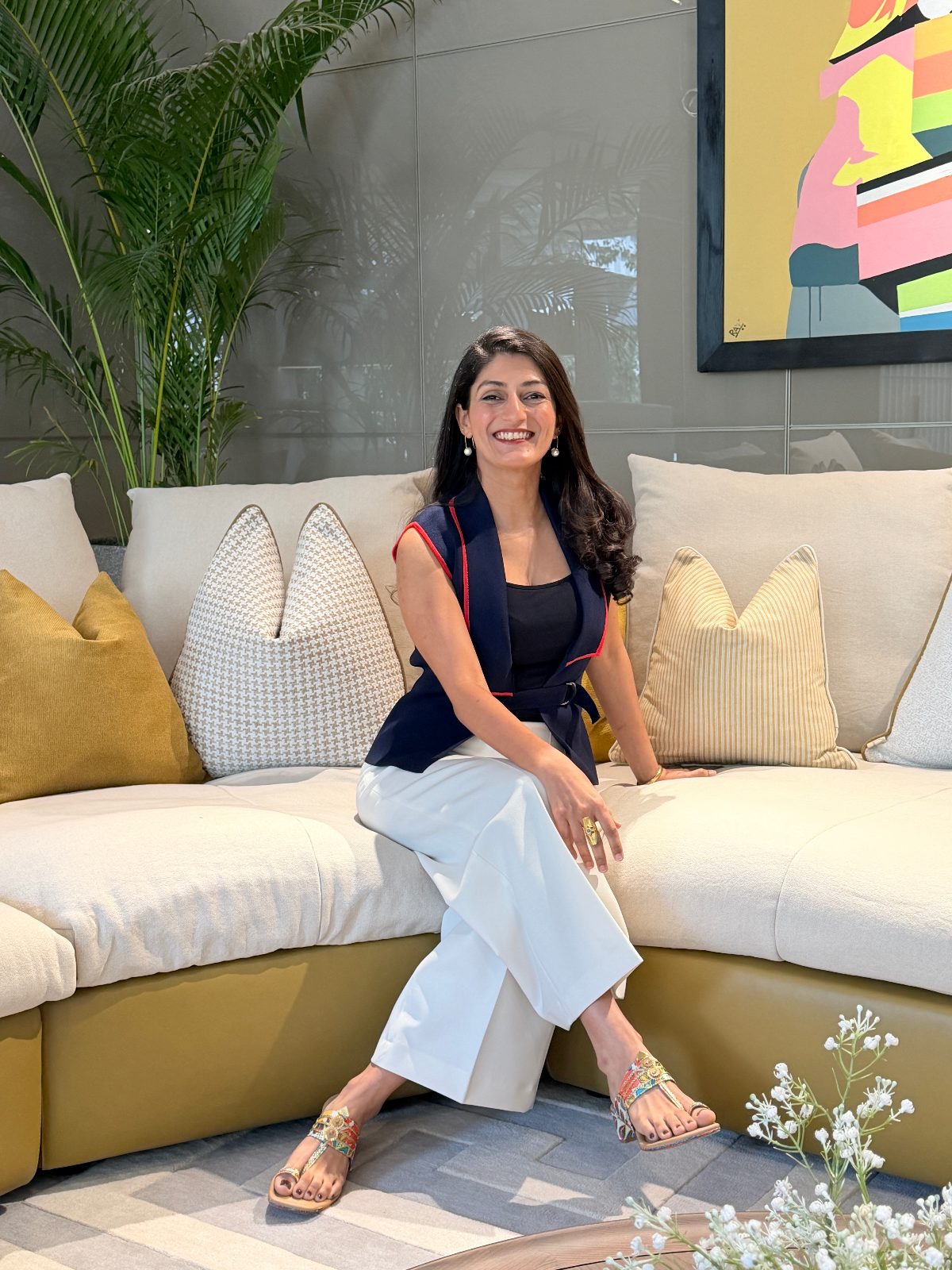 Shreenu Mukherjee, interior designer and co-founder of Atelier Shreenu by The Vrindavan Project, smiling on a curved cream sofa beside a palm.