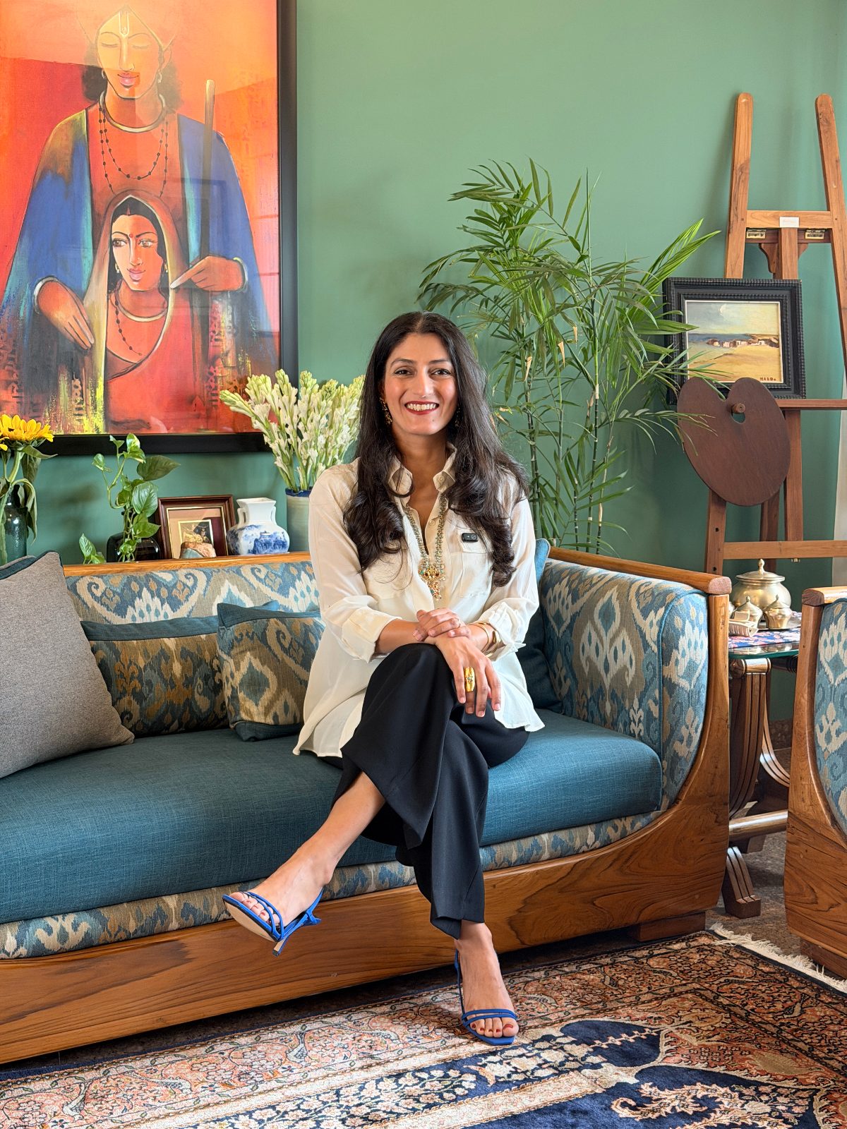 Shreenu Mukherjee, interior designer and co-founder of Atelier Shreenu by The Vrindavan Project, smiling on a teak and ikat sofa in a green room.