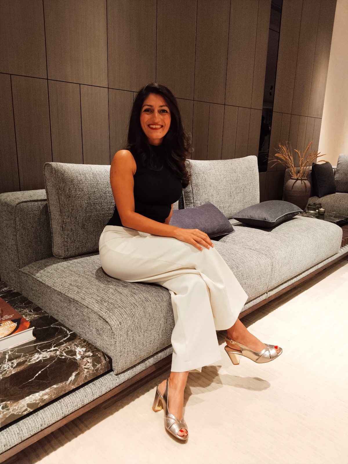 Shreenu Mukherjee smiling on a grey textured sofa in front of walnut wall panelling, with a marble side table beside her.