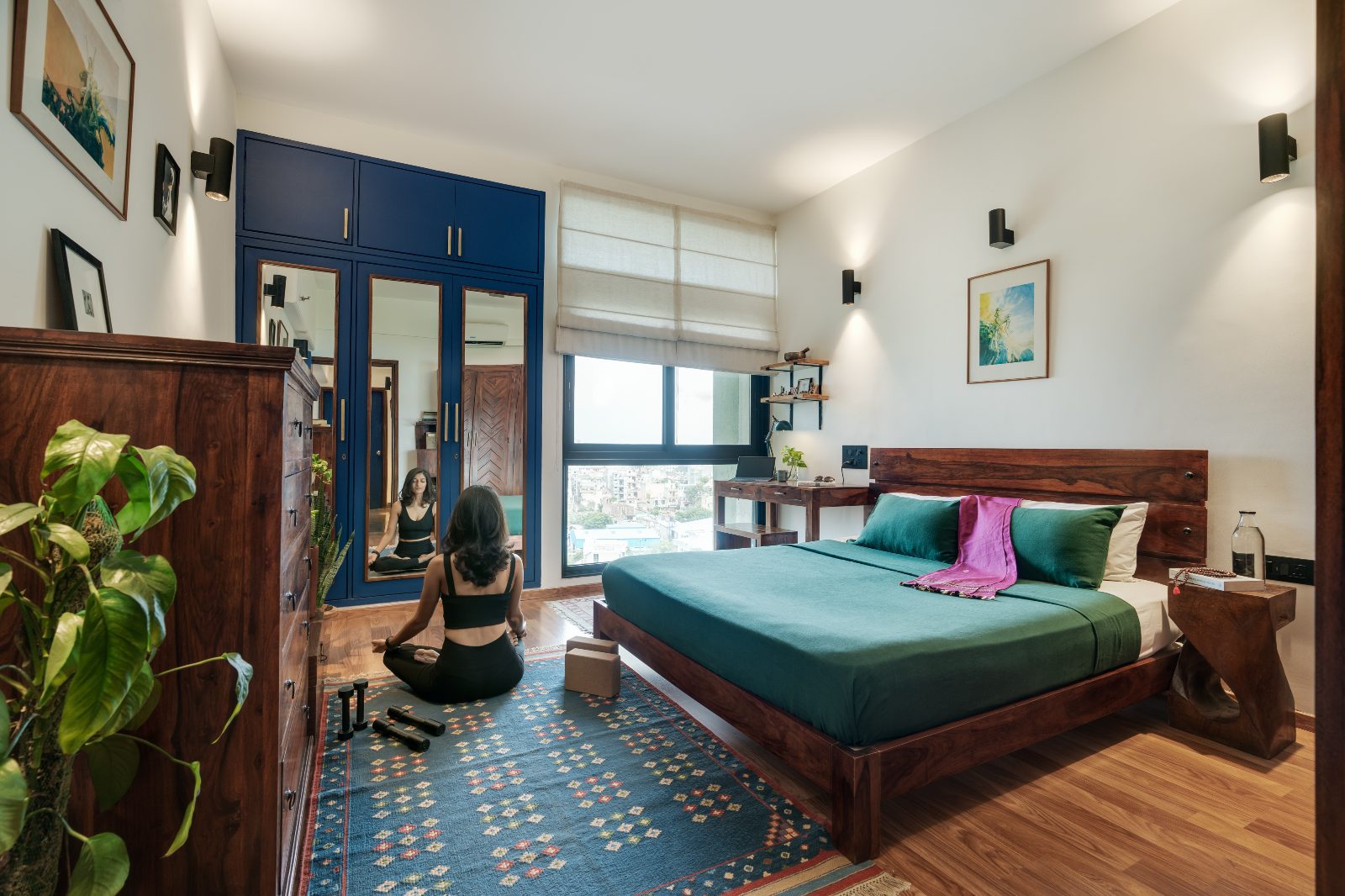 Shreenu Mukherjee meditating on a blue dhurrie in her home office studio, with a teak bed, green linens and a navy mirrored wardrobe.